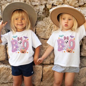 Puede incluir: Dos camisetas blancas de manga corta con un estampado gr&aacute;fico de la palabra BOO en rosa y morado, un fantasma gui&ntilde;ando un ojo, un lazo de lunares, una calabaza naranja sonriente y peque&ntilde;os murci&eacute;lagos. Se llevan con sombreros de ala ancha.