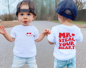 May include: Two young children wearing white t-shirts and blue jeans. One shirt has red sunglasses and the text "Mr. Steal Your Heart". The other shirt has the same text on the back. Both children wear baseball caps.