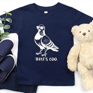 May include: Navy blue toddler t-shirt featuring a white illustration of a pigeon wearing sunglasses. The text "THAT'S COO." is printed below the bird. The shirt is displayed with navy blue shoes, pants, and a teddy bear.