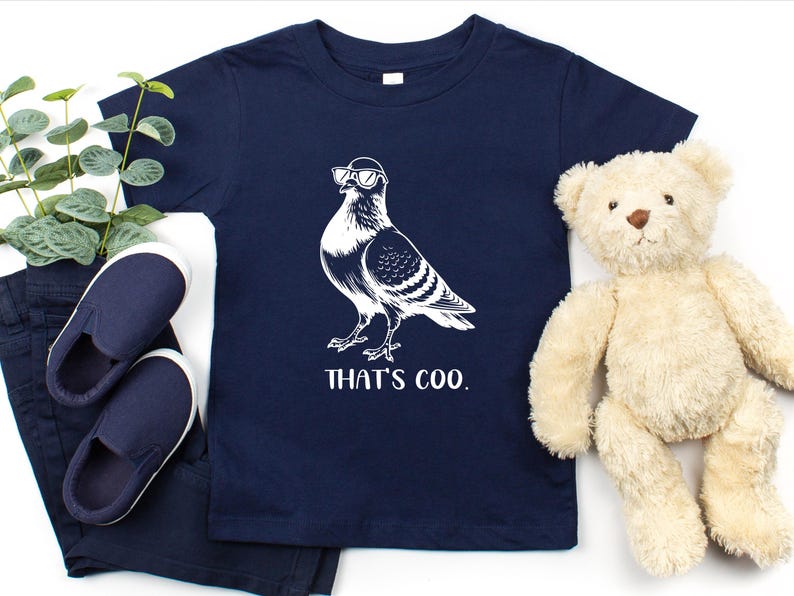 May include: Navy blue toddler t-shirt featuring a white illustration of a pigeon wearing sunglasses. The text "THAT'S COO." is printed below the bird. The shirt is displayed with navy blue shoes, pants, and a teddy bear.