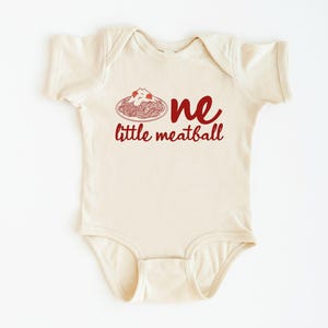 May include: Cream-colored baby onesie with the words "one little meatball" in red script. The design includes a red illustration of spaghetti and meatballs. A cute and playful outfit for a baby.