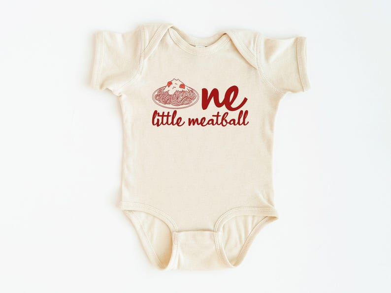 May include: Cream-colored baby onesie with the words "one little meatball" in red script. The design includes a red illustration of spaghetti and meatballs. A cute and playful outfit for a baby.