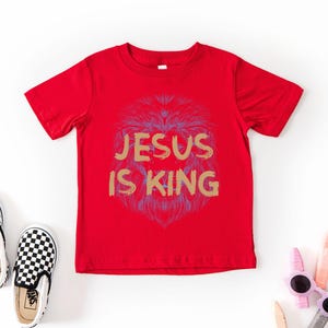 May include: A red t-shirt with the text "JESUS IS KING" in gold lettering and a lion graphic. The image also shows a pair of black and white checkered slip-on shoes and a pair of pink sunglasses.