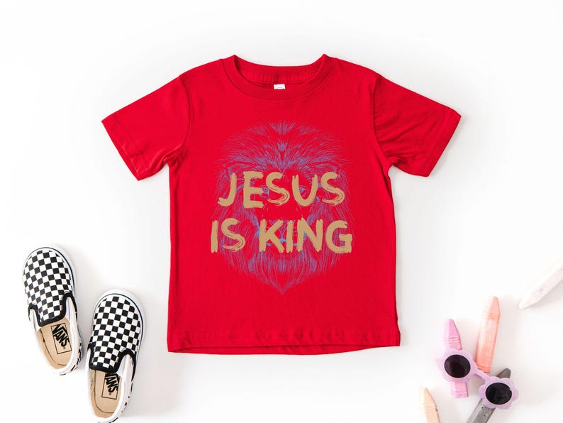 May include: A red t-shirt with the text "JESUS IS KING" in gold lettering and a lion graphic. The image also shows a pair of black and white checkered slip-on shoes and a pair of pink sunglasses.