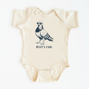 May include: Cream-colored baby bodysuit with a navy blue illustration of a pigeon wearing sunglasses. The text "THAT'S COO." is printed below the bird in navy blue. The bodysuit has short sleeves and a round neckline.