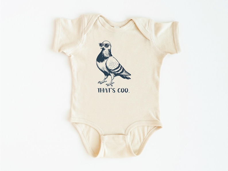 May include: Cream-colored baby bodysuit with a navy blue illustration of a pigeon wearing sunglasses. The text "THAT'S COO." is printed below the bird in navy blue. The bodysuit has short sleeves and a round neckline.