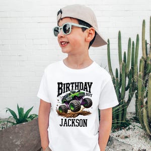 Pode incluir: Camiseta branca com um desenho de monster truck verde e roxo, as palavras "BIRTHDAY BOY" e o nome "JACKSON". A crian&ccedil;a est&aacute; usando &oacute;culos de sol e um bon&eacute; de beisebol.