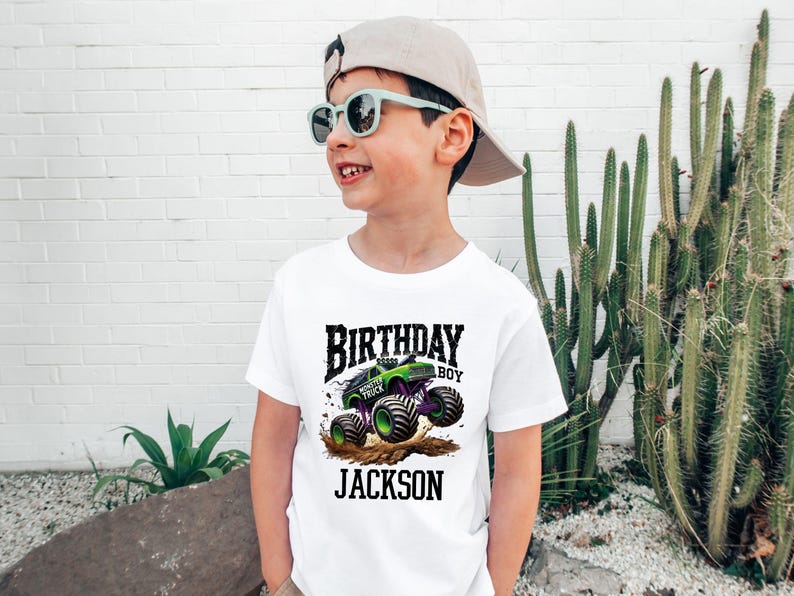 Pode incluir: Camiseta branca com um desenho de monster truck verde e roxo, as palavras "BIRTHDAY BOY" e o nome "JACKSON". A crian&ccedil;a est&aacute; usando &oacute;culos de sol e um bon&eacute; de beisebol.