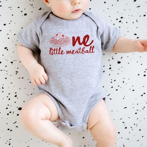 May include: A gray baby onesie with the words "one little meatball" in red script. The design includes a red illustration of spaghetti and meatballs. The baby is lying on a white surface with black splatters.