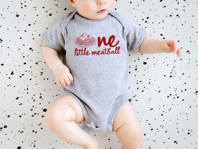 May include: A gray baby onesie with the words "one little meatball" in red script. The design includes a red illustration of spaghetti and meatballs. The baby is lying on a white surface with black splatters.