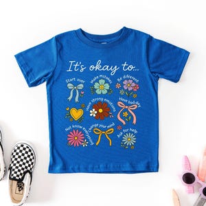 May include: Blue t-shirt with the text "It's okay to..." and colorful illustrations of flowers, bows, and a heart. The shirt is paired with black and white checkered slip-on shoes. The shirt's message encourages self-acceptance.