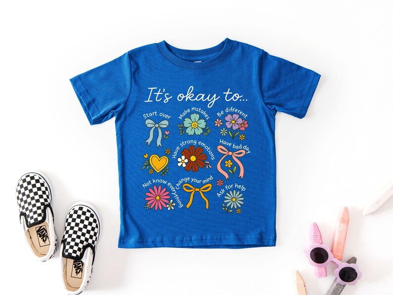 May include: Blue t-shirt with the text "It's okay to..." and colorful illustrations of flowers, bows, and a heart. The shirt is paired with black and white checkered slip-on shoes. The shirt's message encourages self-acceptance.
