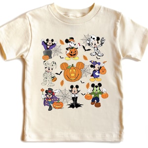 May include: Cream-colored t-shirt featuring a Halloween-themed graphic. The design includes various Mickey Mouse characters dressed in costumes, such as a vampire, mummy, and witch. A large pumpkin with Mickey's face is also featured.