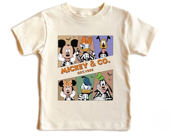 May include: Cream-coloured t-shirt with a graphic of Mickey Mouse, Minnie Mouse, Donald Duck, Daisy Duck, and Goofy as skeletons. The design includes the text "Mickey & Co. EST. 1928" in orange. The shirt has short sleeves.