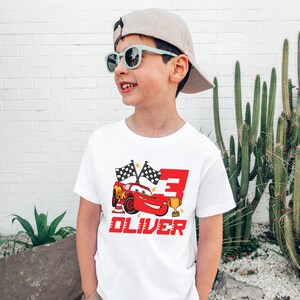 May include: A white t-shirt featuring a red cartoon race car, checkered flags, and the number 3. The name "OLIVER" is printed in red below the car. The boy is wearing sunglasses and a tan cap.