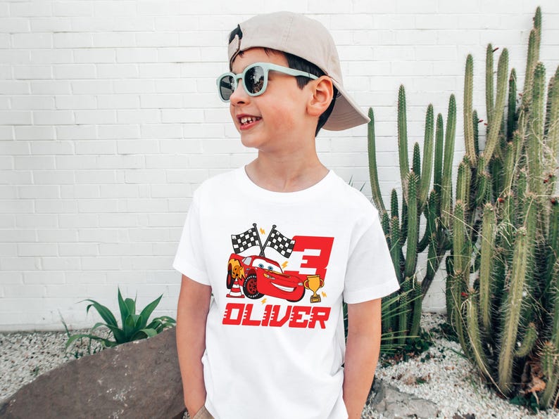 May include: A white t-shirt featuring a red cartoon race car, checkered flags, and the number 3. The name "OLIVER" is printed in red below the car. The boy is wearing sunglasses and a tan cap.