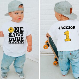 May include: A white t-shirt with the text "ONE HAPPY DUDE" and a smiley face graphic. The shirt also features a checkered pattern at the bottom. The back of the shirt has the name "JACKSON" and the number "1". The child is wearing a gray baseball cap and blue jeans.