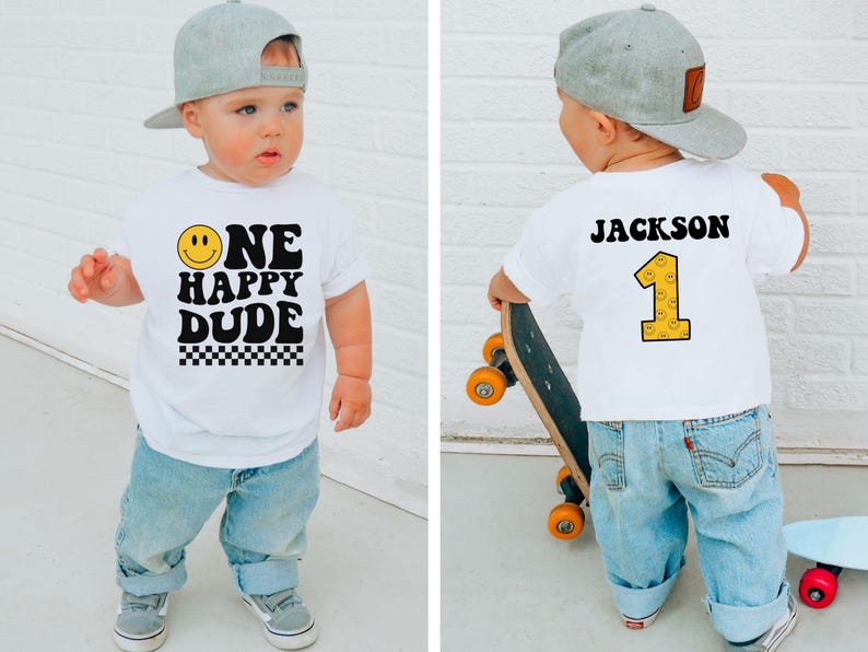 May include: A white t-shirt with the text "ONE HAPPY DUDE" and a smiley face graphic. The shirt also features a checkered pattern at the bottom. The back of the shirt has the name "JACKSON" and the number "1". The child is wearing a gray baseball cap and blue jeans.
