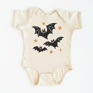 May include: A cream-colored short-sleeve baby bodysuit featuring a graphic print of three black bats and several small orange stars. The garment has a round neckline and snap closures at the bottom, with a textured ink appearance on the bat illustrations.