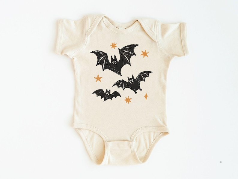 May include: A cream-colored short-sleeve baby bodysuit featuring a graphic print of three black bats and several small orange stars. The garment has a round neckline and snap closures at the bottom, with a textured ink appearance on the bat illustrations.