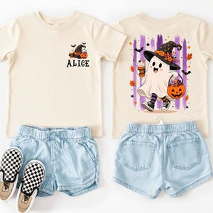 May include: A cream-coloured t-shirt featuring a small ghost graphic with the name Alice on the front left chest, and a large back graphic of a ghost wearing a witch hat, holding a coffee cup and a pumpkin bucket, with purple brush strokes.