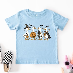 May include: Light blue toddler t-shirt featuring a Halloween-themed graphic. The design includes four cartoon ducks dressed in costumes: a witch, a jack-o'-lantern, a duck holding a black cat, and a vampire duck. Black bats and autumn leaves are also included.
