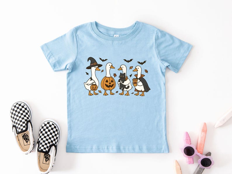 May include: Light blue toddler t-shirt featuring a Halloween-themed graphic. The design includes four cartoon ducks dressed in costumes: a witch, a jack-o'-lantern, a duck holding a black cat, and a vampire duck. Black bats and autumn leaves are also included.