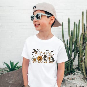 May include: White t-shirt featuring a Halloween-themed design. The graphic depicts four geese dressed in Halloween costumes: a witch, a pumpkin, a cat, and a vampire. Bats are also included in the design.
