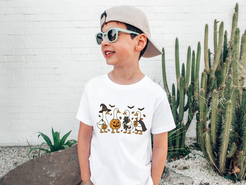 May include: White t-shirt featuring a Halloween-themed design. The graphic depicts four geese dressed in Halloween costumes: a witch, a pumpkin, a cat, and a vampire. Bats are also included in the design.