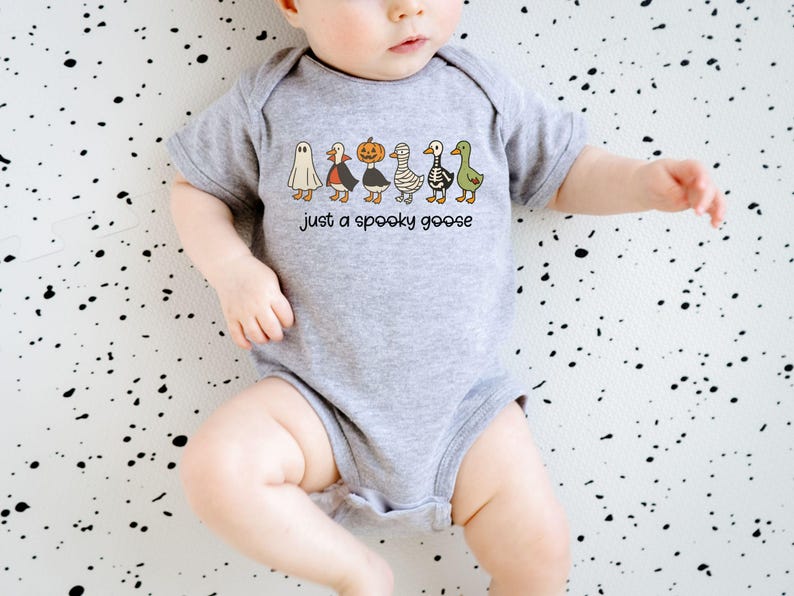 May include: A gray baby onesie with a Halloween-themed graphic. The design features six cartoon geese dressed as a ghost, vampire, pumpkin, mummy, skeleton, and a green goose. Text reads "just a spooky goose".