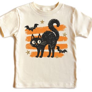 May include: A cream-colored short-sleeve t-shirt featuring a graphic print of a black cat with arched back, two black bats, and several gold-colored stars layered over three horizontal orange brushstroke stripes on the front center.