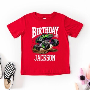 Pode incluir: Uma camiseta vermelha com um gr&aacute;fico de um monster truck verde e roxo em um monte de terra. O texto branco diz "BIRTHDAY BOY JACKSON". Sapatos slip-on xadrez em preto e branco est&atilde;o &agrave; esquerda.