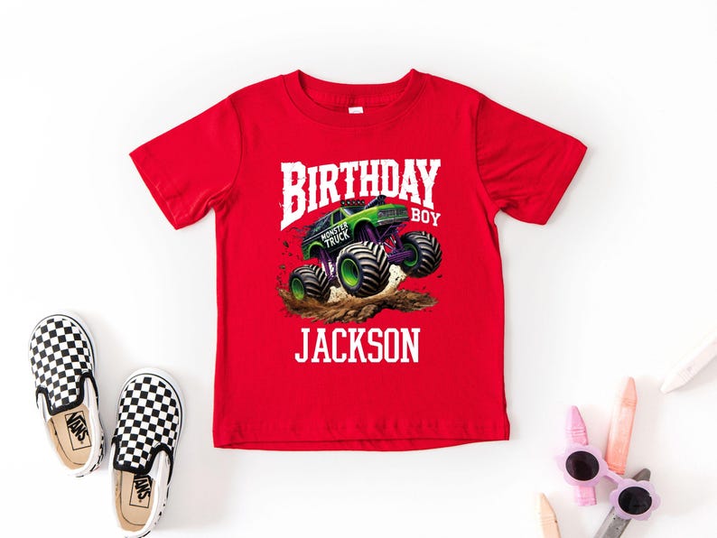 Pode incluir: Uma camiseta vermelha com um gr&aacute;fico de um monster truck verde e roxo em um monte de terra. O texto branco diz "BIRTHDAY BOY JACKSON". Sapatos slip-on xadrez em preto e branco est&atilde;o &agrave; esquerda.