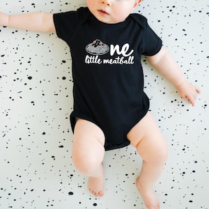 May include: A black baby onesie with the words "one little meatball" in white script. The design includes a graphic of spaghetti and meatballs. The baby is lying on a white surface with black splatters.