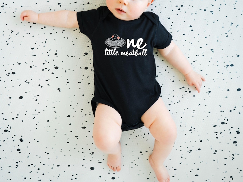 May include: A black baby onesie with the words "one little meatball" in white script. The design includes a graphic of spaghetti and meatballs. The baby is lying on a white surface with black splatters.