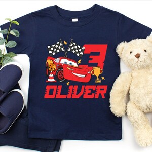 May include: Navy blue t-shirt featuring a red Lightning McQueen graphic with the number 3, checkered flags, and a trophy. The name "OLIVER" is printed in red below the graphic. Includes navy blue shoes, pants, and a teddy bear.