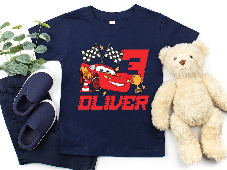 May include: Navy blue t-shirt featuring a red Lightning McQueen graphic with the number 3, checkered flags, and a trophy. The name "OLIVER" is printed in red below the graphic. Includes navy blue shoes, pants, and a teddy bear.