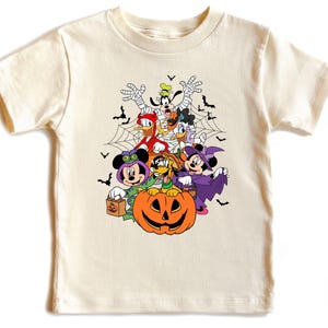 Op de afbeelding: Cr&egrave;mekleurig T-shirt met een Halloween-thema. Het ontwerp bevat stripfiguren in kostuums, een pompoen, vleermuizen en een spinnenweb. De personages zijn Mickey Mouse, Minnie Mouse, Donald Duck, Goofy en Daisy Duck.