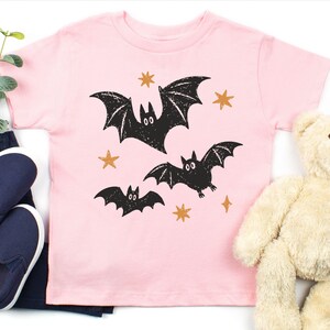 May include: A light pink short-sleeve t-shirt featuring a graphic print of three black bats in flight surrounded by small orange stars. The design has a textured, distressed appearance, and the shirt is made of a solid-colored cotton fabric.