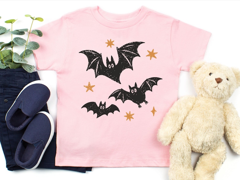 May include: A light pink short-sleeve t-shirt featuring a graphic print of three black bats in flight surrounded by small orange stars. The design has a textured, distressed appearance, and the shirt is made of a solid-colored cotton fabric.