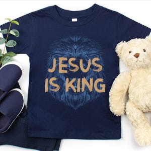 May include: Navy blue toddler t-shirt with the text "JESUS IS KING" in gold lettering over a blue lion's head design. The image includes navy blue shoes, pants, and a teddy bear.