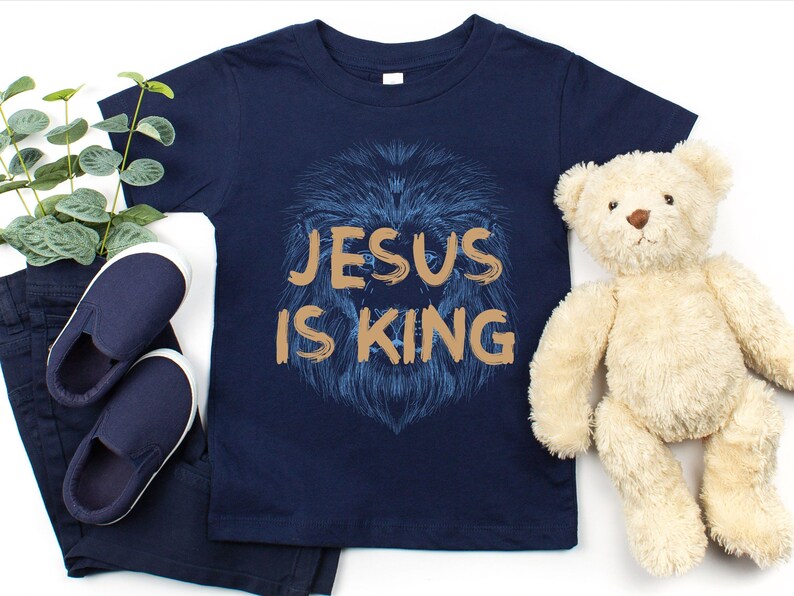 May include: Navy blue toddler t-shirt with the text "JESUS IS KING" in gold lettering over a blue lion's head design. The image includes navy blue shoes, pants, and a teddy bear.