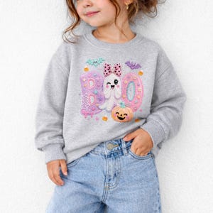 Puede incluir: Una sudadera gris jaspeado de manga larga con un estampado gr&aacute;fico de la palabra BOO en rosa y morado, un fantasma blanco con un lazo rosa de lunares, una calabaza naranja sonriente, murci&eacute;lagos peque&ntilde;os, estrellas y caramelos.