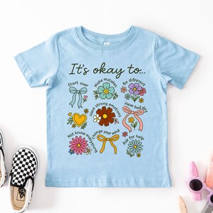 May include: A light blue t-shirt with the phrase "It's okay to..." and various floral and bow illustrations. The shirt includes phrases like "Start over", "Make mistakes", and "Have a bad day". A pair of black and white checkered slip-on shoes are next to the shirt.