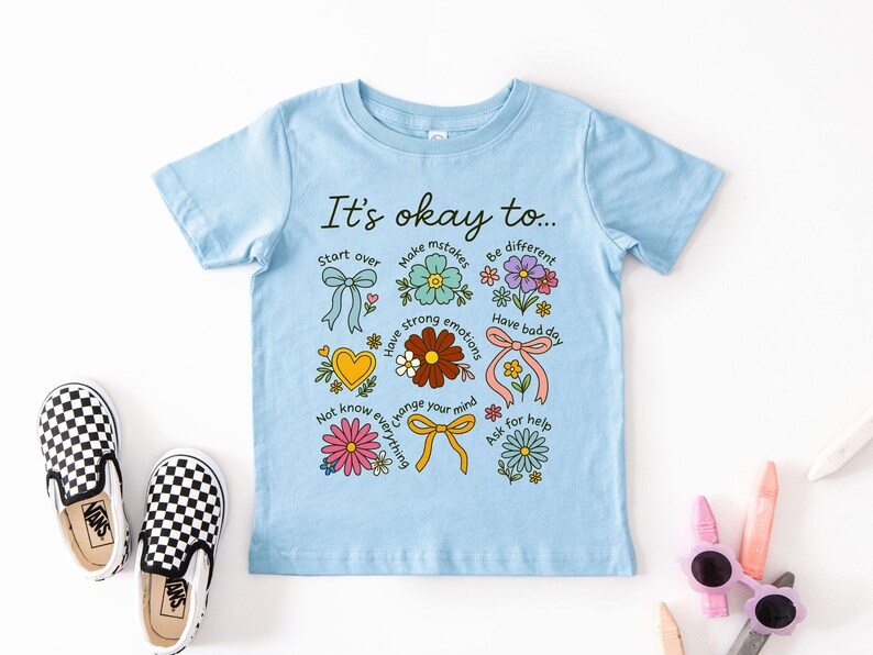 May include: A light blue t-shirt with the phrase "It's okay to..." and various floral and bow illustrations. The shirt includes phrases like "Start over", "Make mistakes", and "Have a bad day". A pair of black and white checkered slip-on shoes are next to the shirt.