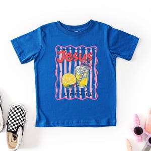May include: A royal blue t-shirt featuring a graphic of a lemonade drink with a lemon slice and the text "Jesus Lemonade" in a retro style. The shirt is surrounded by a pink and white striped border. Black and white checkered slip-on shoes are on the left.