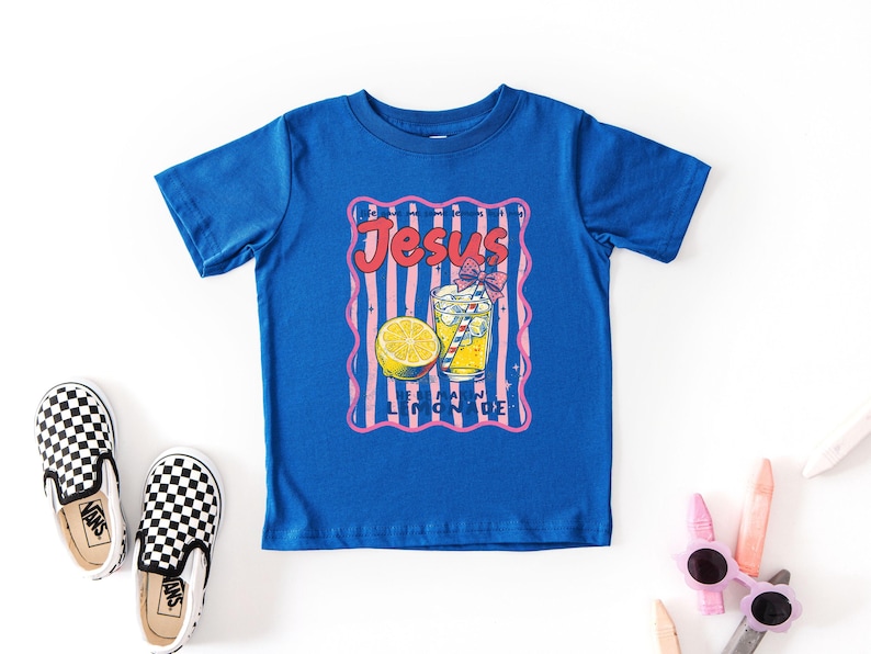 May include: A royal blue t-shirt featuring a graphic of a lemonade drink with a lemon slice and the text "Jesus Lemonade" in a retro style. The shirt is surrounded by a pink and white striped border. Black and white checkered slip-on shoes are on the left.