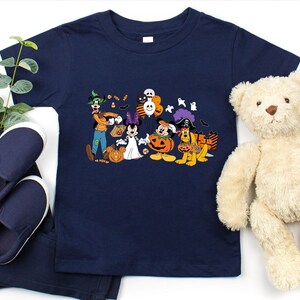 May include: Navy blue t-shirt featuring a Halloween-themed graphic with Disney characters dressed in costumes. The design includes Mickey Mouse, Minnie Mouse, Goofy, and Pluto. The shirt is paired with navy blue pants and shoes, and a teddy bear.
