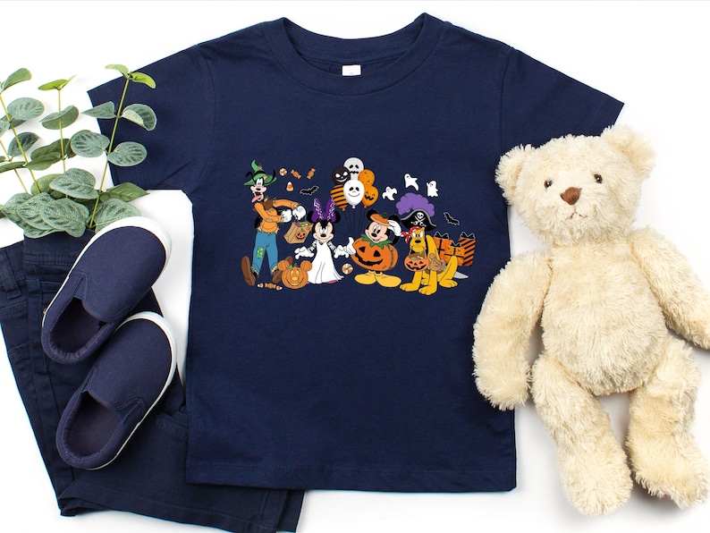 May include: Navy blue t-shirt featuring a Halloween-themed graphic with Disney characters dressed in costumes. The design includes Mickey Mouse, Minnie Mouse, Goofy, and Pluto. The shirt is paired with navy blue pants and shoes, and a teddy bear.