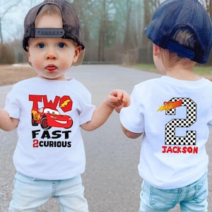 May include: Two white t-shirts with a Cars theme. One shirt features Lightning McQueen and the text "TWO FAST 2 CURIOUS". The other shirt has a checkered number 2 with the name "JACKSON". Both shirts are for toddlers.